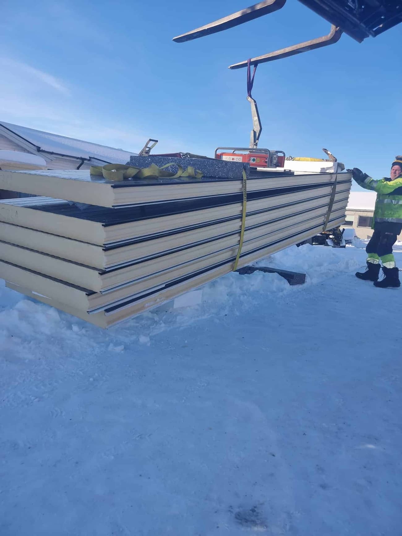 En truck løfter en stabel sandwichelementer på et snødekt anlegg under klar himmel